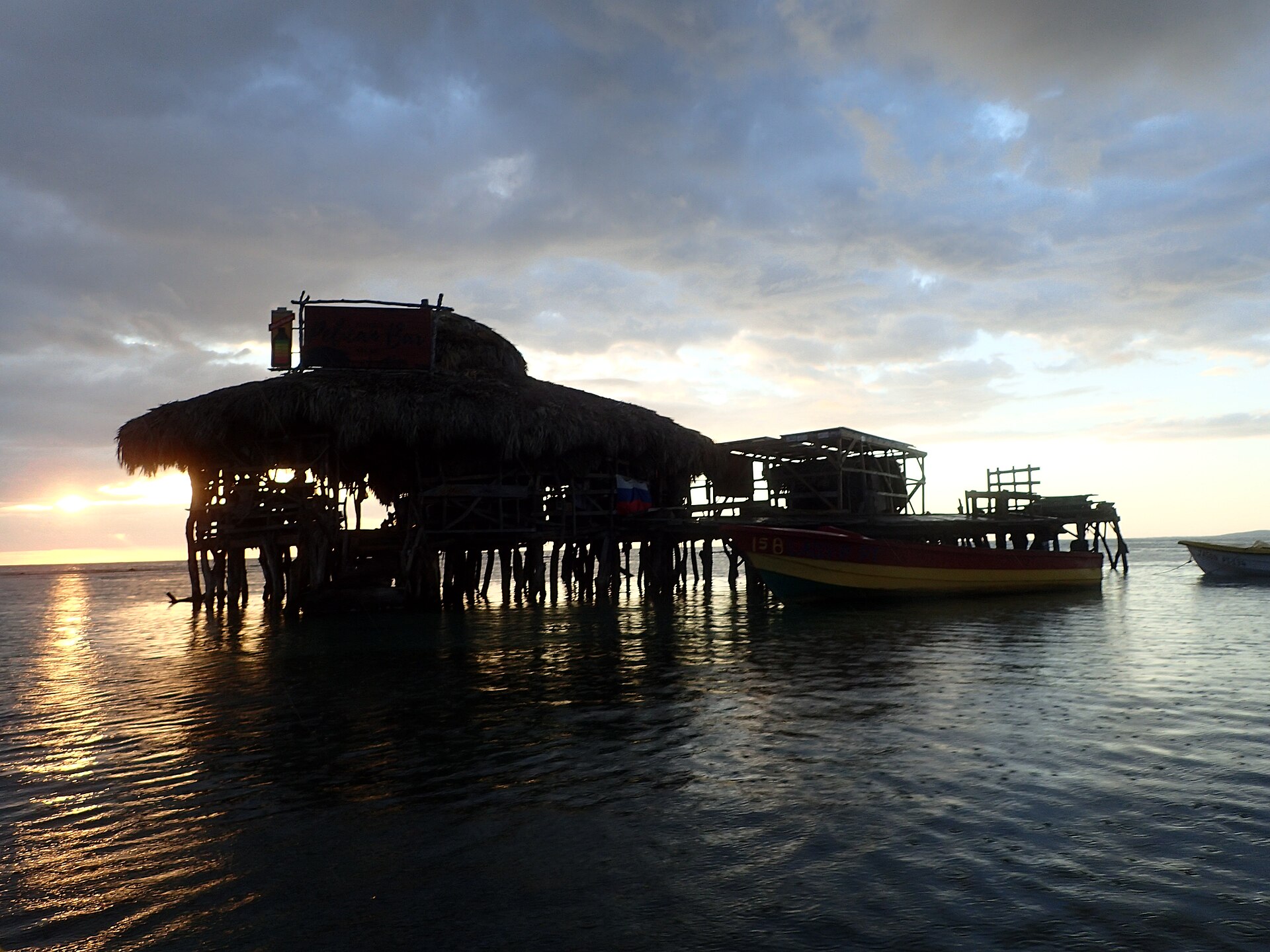 Pelican Bar, a thatched wooden bar on stilts in the sea off Treasure Beach, St Elizabeth, at sunset with a fishing boat alongside